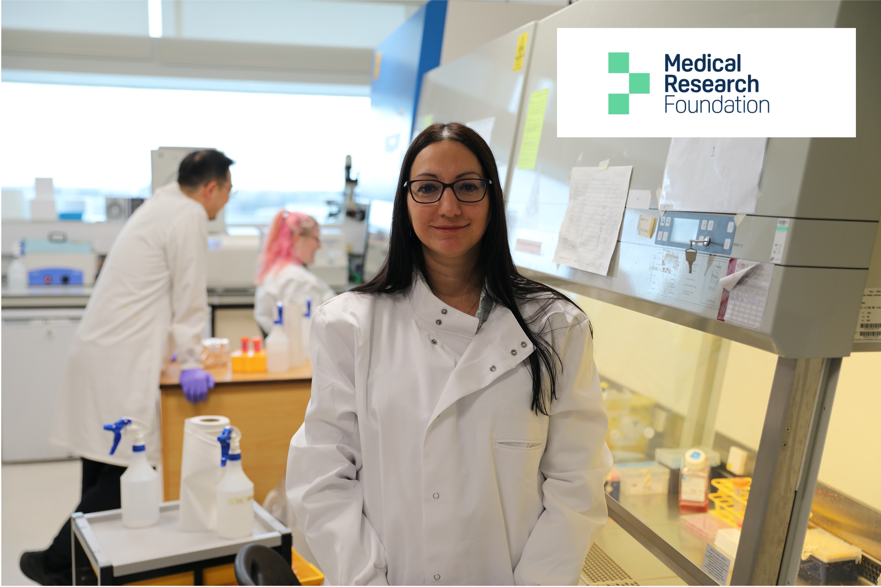 A woman wearing a lab coat, standing in front of two PhD students in the lab